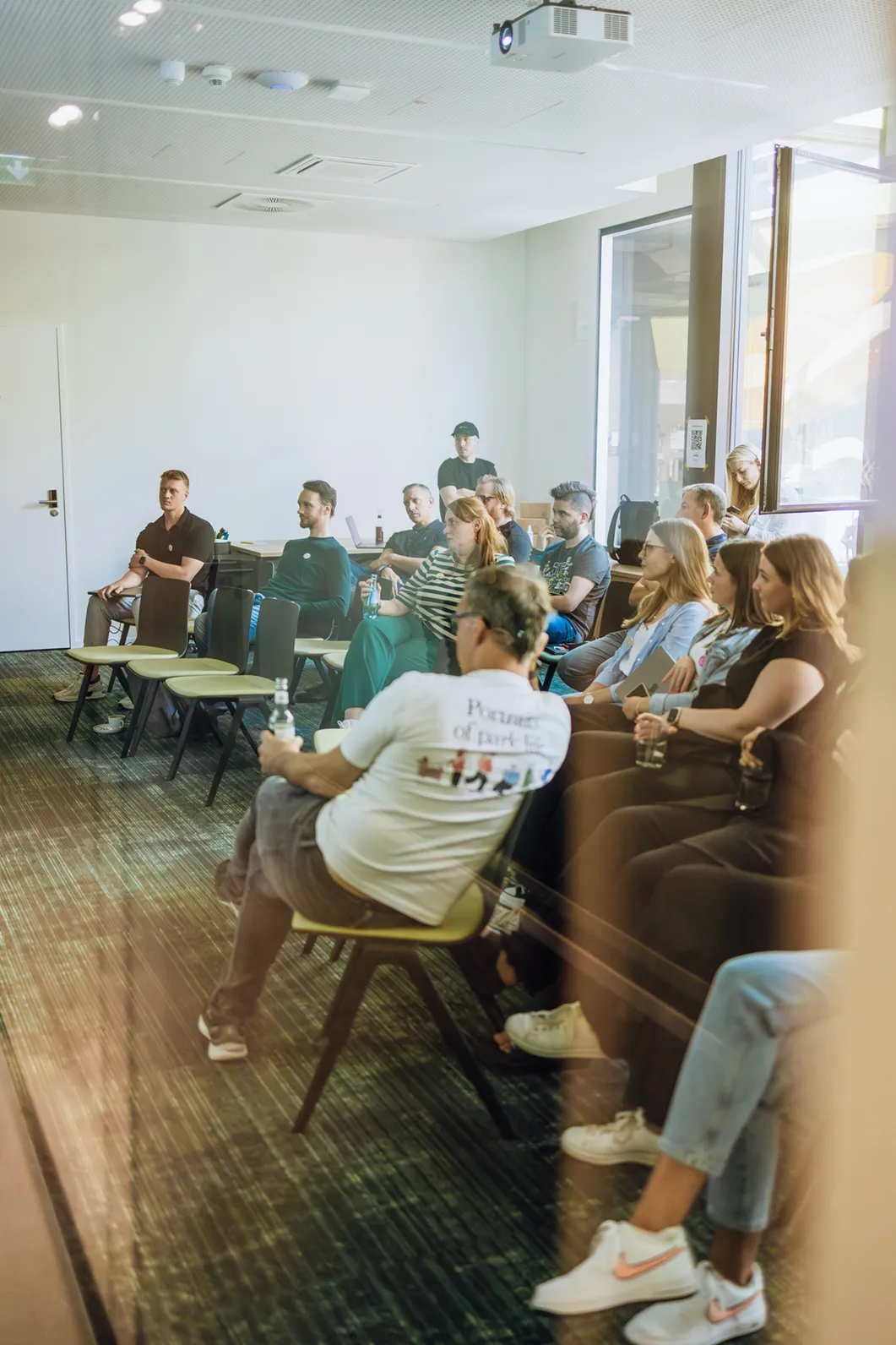 A group of people seated in a conference room, attentively listening to a presentation. The room has a projector on the ceiling and large windows on one side.