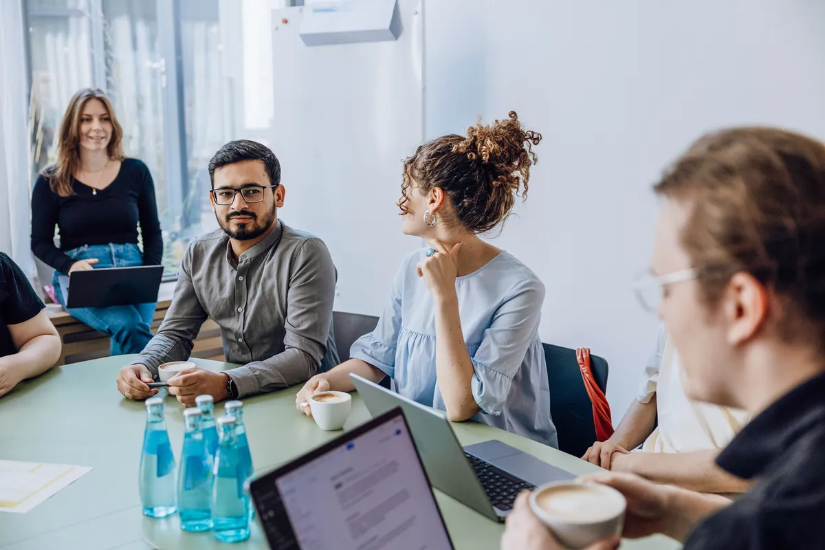 A group of people sitting around a table in a modern office setting, engaged in a casual meeting. They have laptops and coffee cups in front of them, and there are bottles of water on the table. One person is standing and smiling, while others are seated and conversing.