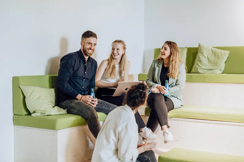 A group of four people sitting on green cushioned benches, smiling and holding drinks, with a laptop open in front of them.
