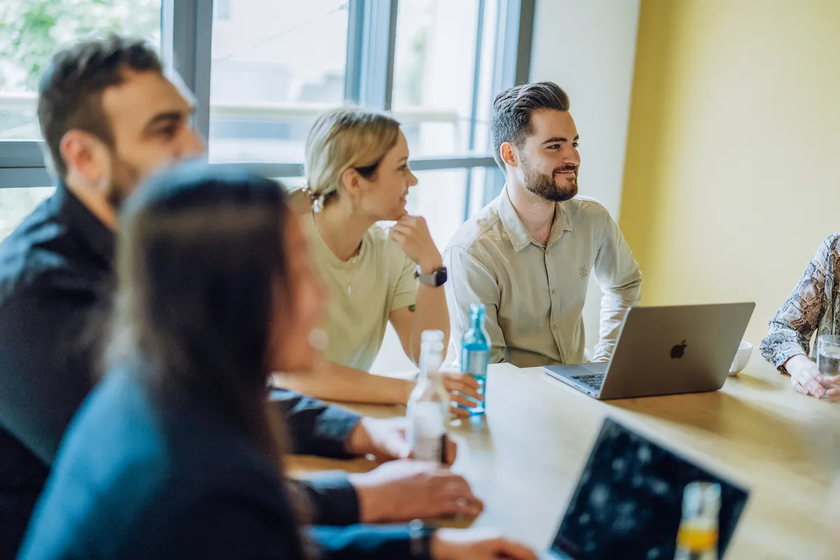A group of people sitting around a table with laptops and drinks, engaged in a discussion in a bright room with large windows.