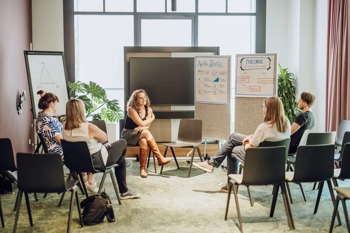 A group of people sitting in a circle in a bright room, engaged in a discussion. There are flipcharts with diagrams and notes in the background.
