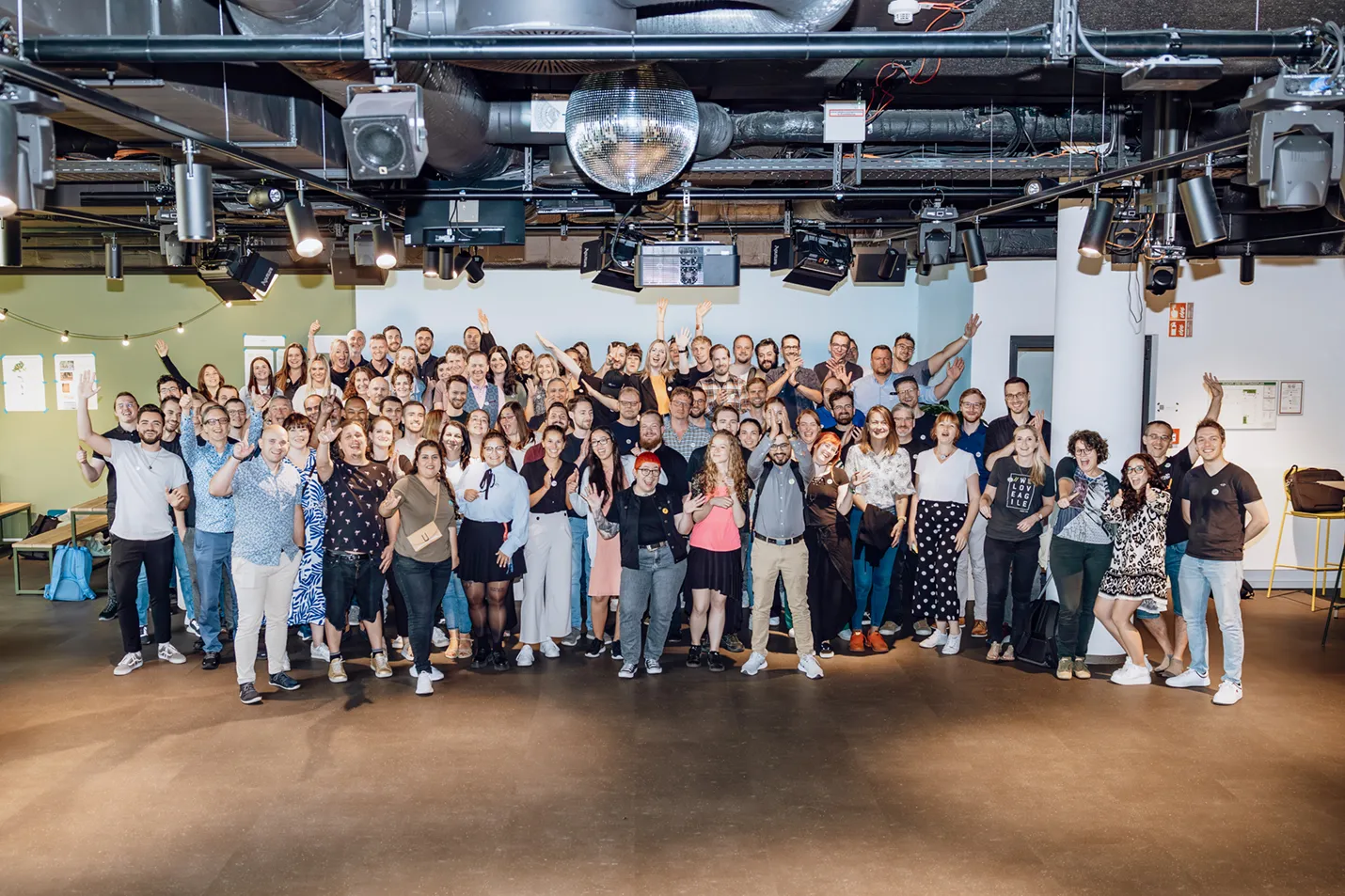 A large group of people posing together indoors, smiling and waving at the camera. The setting appears to be a modern office or event space with a disco ball on the ceiling.