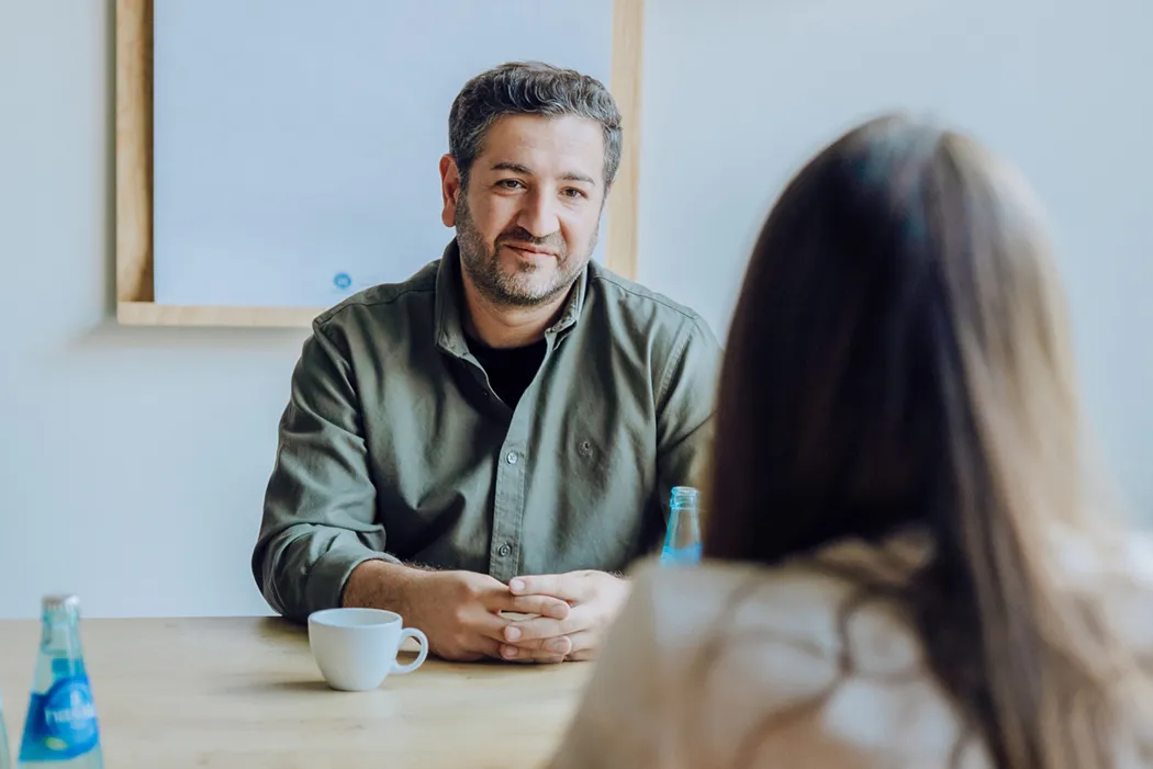 A man in a green shirt sits at a table with a cup of coffee and a bottle of water, facing a woman with long hair.