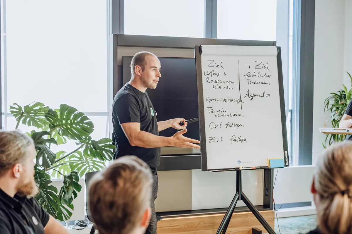 A man presenting in an office setting, standing next to a flipchart with handwritten notes. Several people are seated, listening attentively. Large windows and plants are visible in the background.