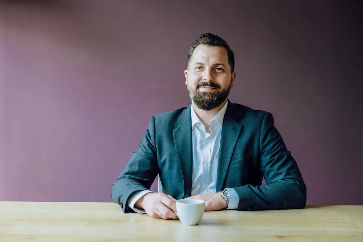 Man in a suit sitting at a table with a cup of coffee, smiling against a purple background.