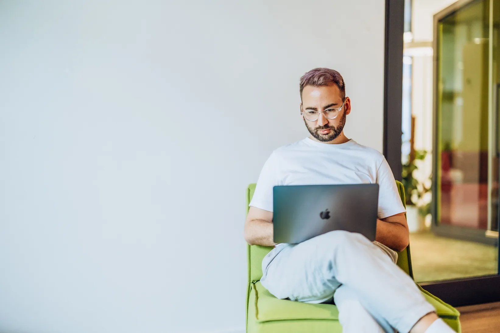 A man sitting on a green chair, focused on working on his laptop in a modern office setting.