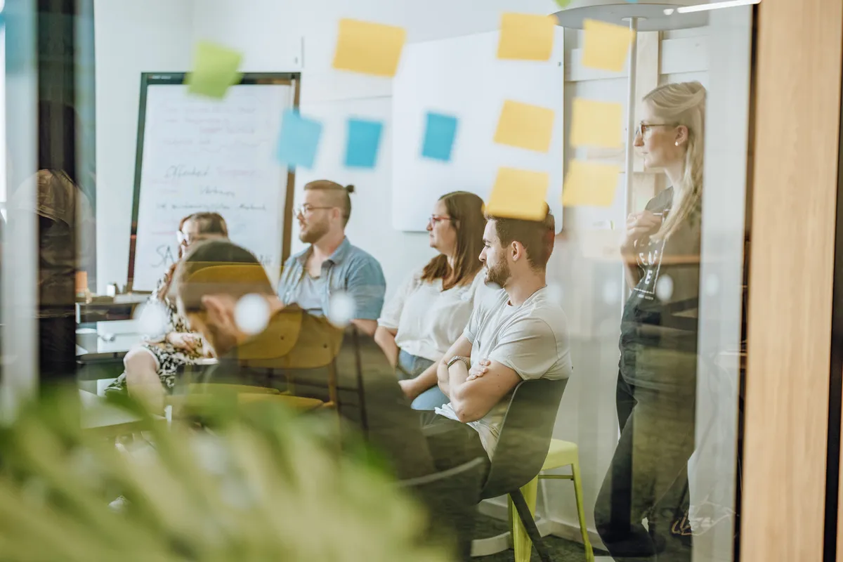 A group of people sitting and standing in a meeting room, viewed through a glass wall with sticky notes attached.