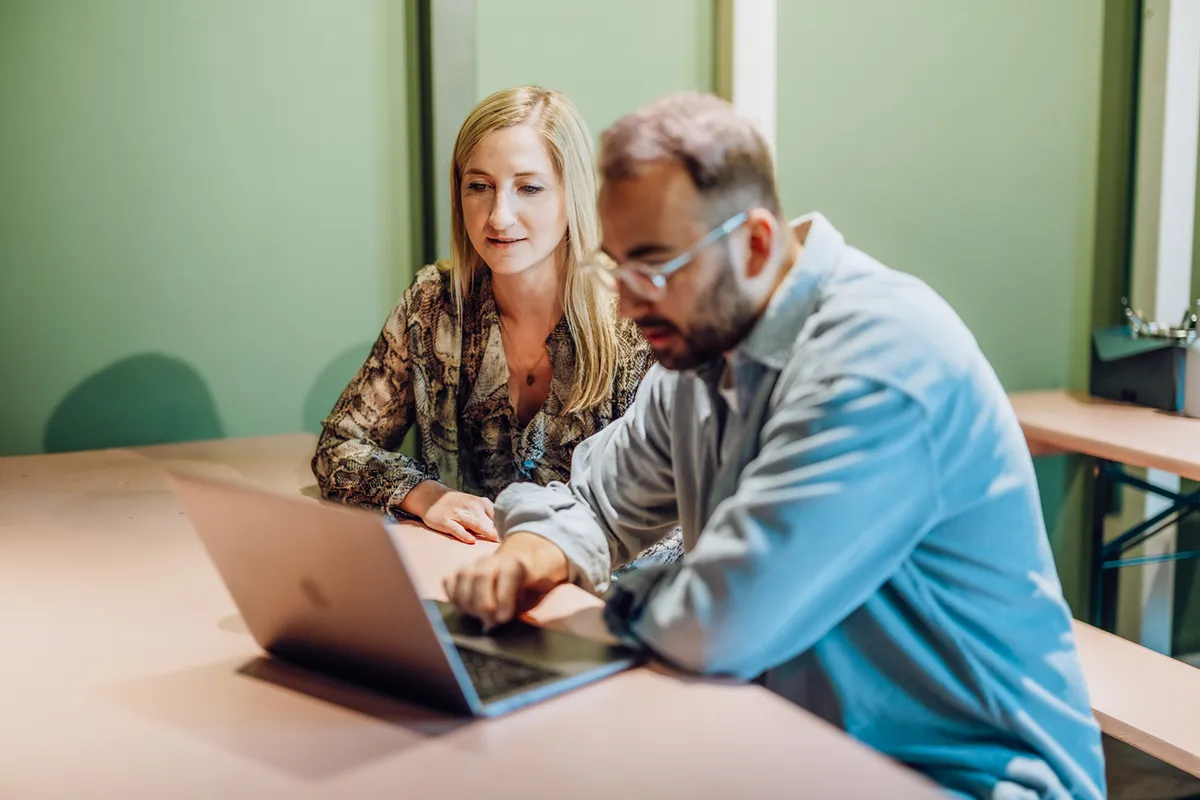 Two people sitting at a table looking at a laptop screen, engaged in a discussion.
