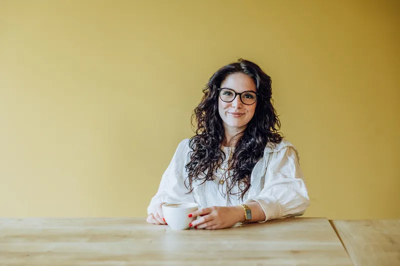 Woman with glasses holding a coffee cup, sitting at a wooden table against a yellow background.