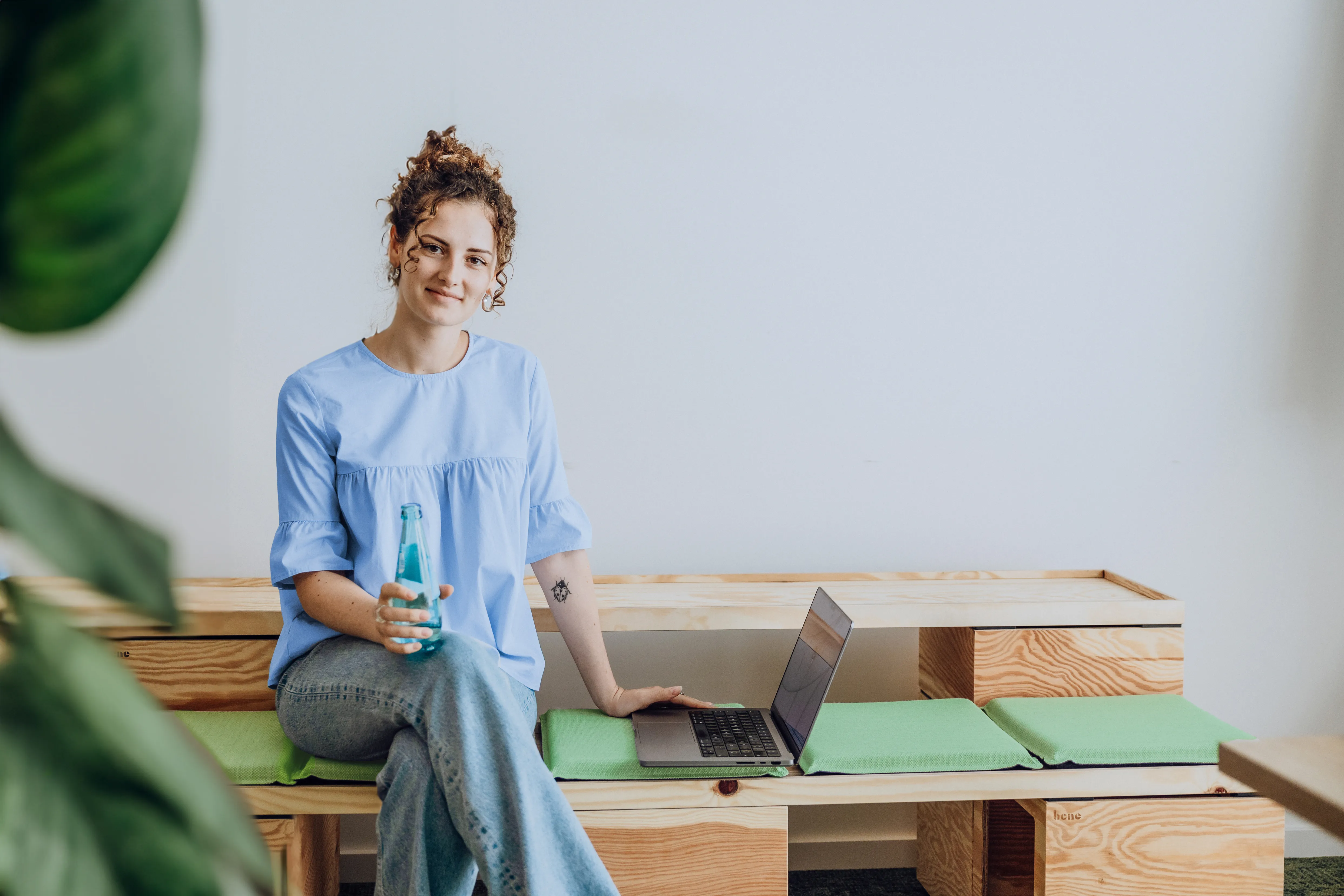 A woman in a blue shirt sitting on a wooden bench with a laptop and holding a bottle, smiling at the camera.