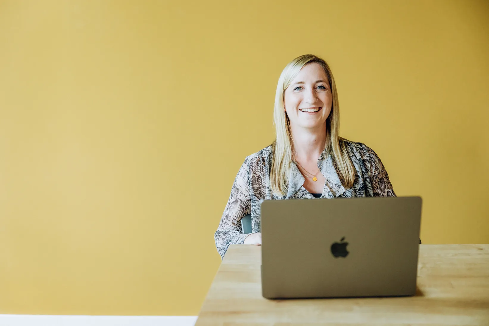 Woman smiling while sitting at a table with a laptop in front of a yellow wall.
