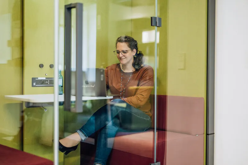 Woman sitting in a small glass-walled office booth, working on a laptop with a smile. She is wearing glasses and a brown top, and there is a bottle of water on the table.